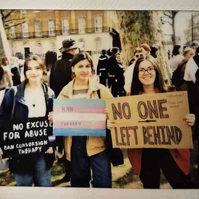 Three individuals stand holding signs advocating for banning conversion therapy and inclusivity during a protest. One sign says "NO EXCUSE FOR ABUSE BAN CONVERSION THERAPY", one says "BAN CONVERSION THERAPY" and the third says "NO ONE LEFT BEHIND".