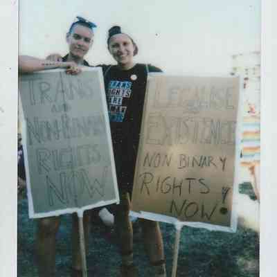 Two people hold signs which say "Trans and Non-binary Rights Now" and "Legalise My Existence Non-binary Rights Now".