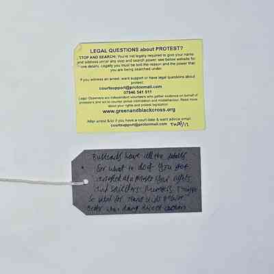 A yellow "Bust Card" from Green and Black Cross detailing a persons rights around Stop and Search and contact details for further advice. With a brown, handwritten luggage tag.