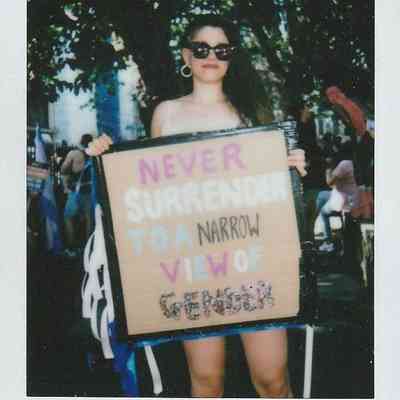 A person is holding a sign that reads "Never Surrender to a Narrow View of Gender".