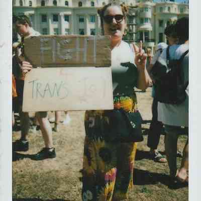 A person wearing sunglasses holds a sign saying "Fight for Trans Joy" in a sunny outdoor gathering.