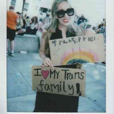 A person smiles, holding signs supporting the trans community, one says "Trans Pride" with a rainbow drawn on and the other says "I <3 My Trans Family".