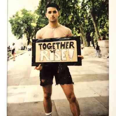 A person stands on a street holding a sign that reads, "TOGETHER WE RISE!"