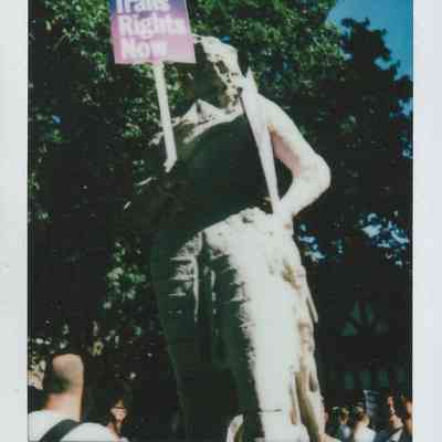 A statue holds a sign reading "Trans Rights Now" at a gathering outdoors under a bright blue sky