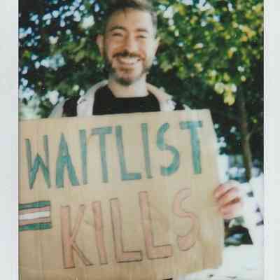 A smiling person holds a sign reading "WAITLIST KILLS" in a sunny park setting.