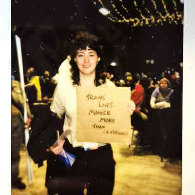 A person holds a sign reading "Trans lives matter more than cis feelings" at a crowded event.