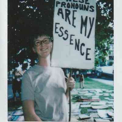A person holds a sign saying, "These pronouns are my essence," while standing outdoors under trees