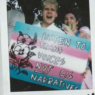 Two individuals hold a colorful sign advocating for listening to trans voices over cis narratives.