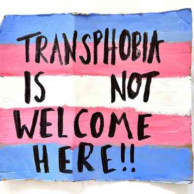 The sign displays "Transphobia is not welcome here" on a flag background.