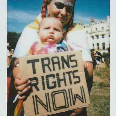 A person with colorful makeup holds a baby and a "Trans Rights Now".