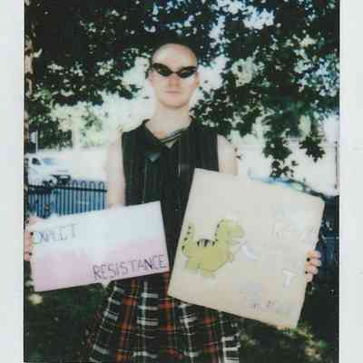 A person in sunglasses holds two signs, "Expect Resistance" and a dinosaur drawing, outdoors.