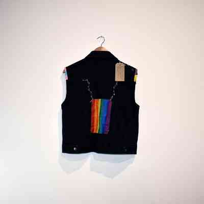 A dark vest with rainbow fabric hangs against a plain white wall.