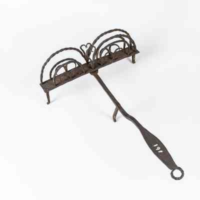 The image shows an antique cooking tool with an ornate design and a long twisted handle. / L'image montre un outil de cuisine antique avec un design orné et un long manche torsadé.