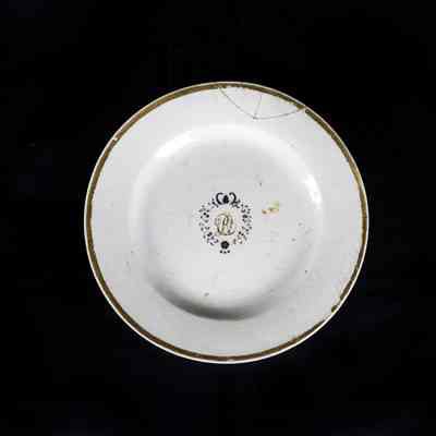 A white ceramic plate with a decorative emblem and gold rim sits on a dark background. / Une assiette en céramique blanche avec un emblème décoratif et un bord doré repose sur un fond noir.