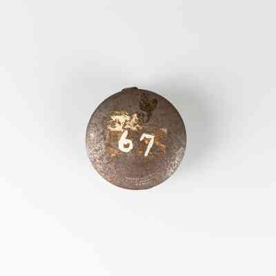 A rusty, round, metallic box with the number 67 painted in it, is centered on a plain white background / Une boîte métallique ronde et rouillée, sur laquelle est peint le chiffre 67, est centrée sur un fond blanc uni.