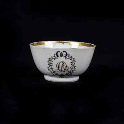 A small white ceramic cup features a decorative design with gold trim, set against a black background. / Une petite tasse en céramique blanche présente un motif décoratif avec une bordure dorée, sur un fond noir.