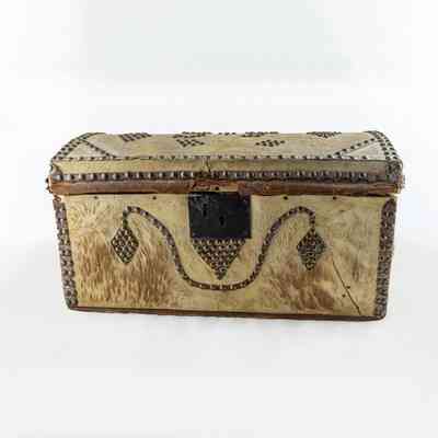 The image shows an ornate but worn beige coloured chest with intricate metal decorations on a neutral background. / L'image montre un coffre beige orné mais usé, avec des décorations métalliques complexes sur un fond neutre.