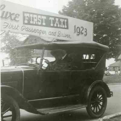 Man Driving a Passenger Past DeLuxe First Taxi Sign
