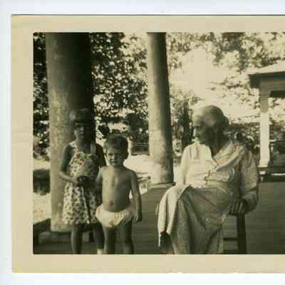 Photograph of an old woman sitting on a porch besides a young girl and young boy