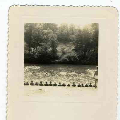 Photograph of people swimming in a pond and others watching from the banks