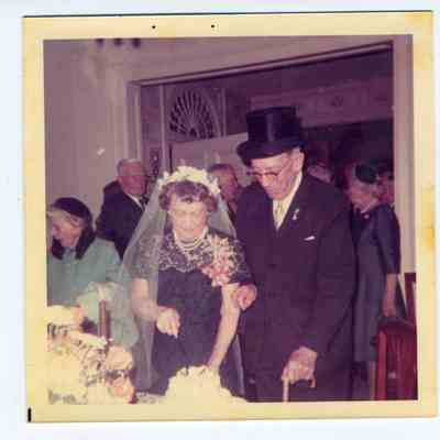 Photograph of an older couple and guests, bride cutting a cake