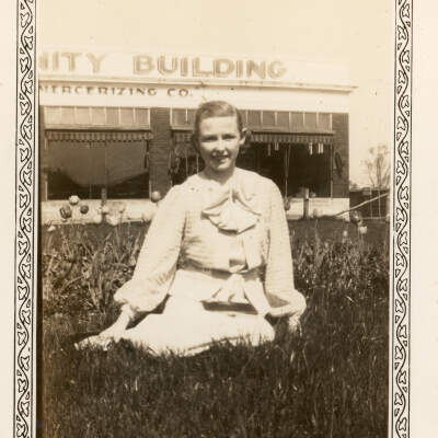 2022.098.002 Selection: Black and white photograph of a young woman seated on the grass amongst tulips in front of the Dixie Mercerizing Company building