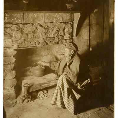 2022.057.076 Recto: Black and white photograph of an unknown woman in front of a stone fireplace