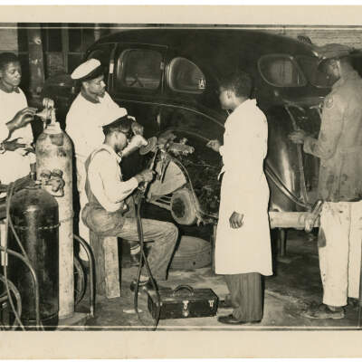 2022.024.016 Recto: Black and white photograph showing a Boone Higgins Trade School student using a torch to make repairs on a car