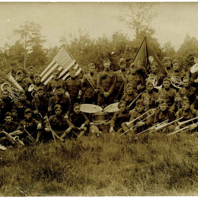 2020.576.002 Recto: Photographic postcard featuring a WWI era marching band group photograph. There are 36 men posing with their instruments with a drum set in the middle and flags at the rear.