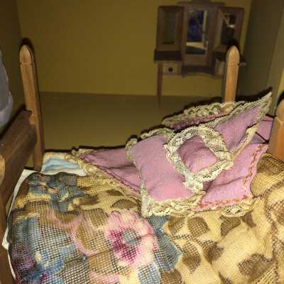 Bed and dresser with three mirrors: The bed has the following fabric pieces: 
- mattress 
- blue flannel pad
- sheet (handkerchief)
- quilted duvet (multicolored fabric with floral pattern)
- bedspread (pink with lace edging on 3 sides)
- doily/pink cloth square edged with lace
- large oblong pink cushion with lace edging
- small oblong cushion with lace edging

Dresser has movable side mirrors and drawers.
