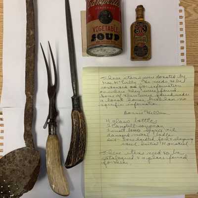Mac McEvilly donated kitchen items: Specifically, from left to right these items include:
- A severely corroded straining ladle (likely $0 value);
- A real antler handled antique serving fork and sharpening steel (perhaps a $50 value);
- A vintage Cambell's Soup can (perhaps a $0 to $5 value given its condition), and;
- A partially full Signet Oil bottle (perhaps a $2 to $7 value given its condition).