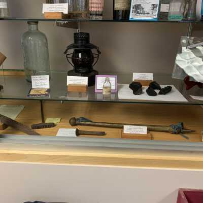 Various Artifacts and Photos Display - Right Side Close Up 1 Bottom Shelves: Various Artifacts and Photos Display - Right Side Close Up 1 Bottom Shelves.  Includes a large Sand Lake Springs Bottle, railroad lantern and spike, antique glass pane, glass beads, J. K. Averill branding iron, charcoal display, and Rebekah Lodge official door symbol.