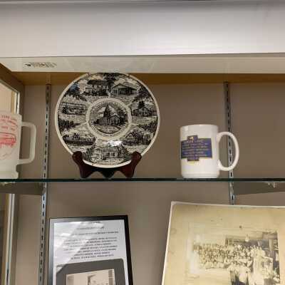 Various Artifacts and Photos Display - Left Side Close Up Top Shelf: Various Artifacts and Photos Display - Left Side Close Up Top Shelf.  Includes a West Sand Lake Fire Department mug, Sand Lake US Bicentennial Plate, Burden Lake Historic marker mug, and Sand Lake Springs bottle.  