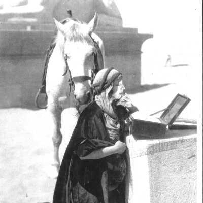 Mirium played by Estelle Taylor fixing makeup in small mirror. White horse stands in the background.