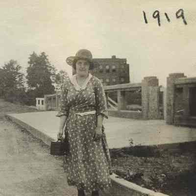 c.1919, an unidentified woman stands in front of the Lee Road and Van Aken Boulevard Rapid Transit entrance. Warrensville West Cemetery is visible in the background to the left, and East View School is in the background to the right.