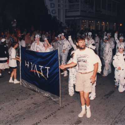 Two unknown people holing a sign that reads the Hyatt with unknown people dressed up walking behind them during the parade.