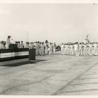 A group of unknown people in uniform standing while one speaks at the podium