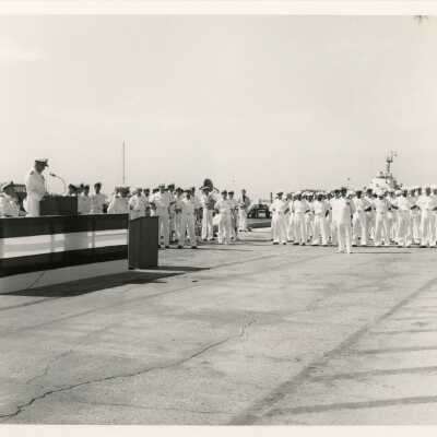 A group of unknown people in uniform standing while one speaks at the podium