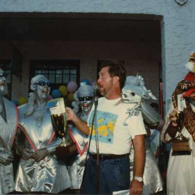 Unknown man talking at a microphone with people dressed up standing around him.