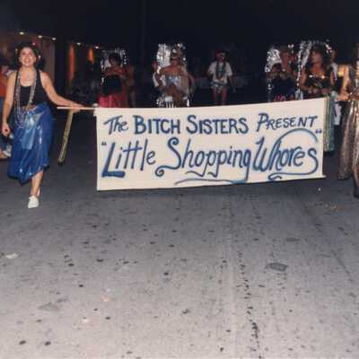 Unknown people dressed up walking down the street holding a sign that reads The Bitch sisters present Little Shopping Whores.
