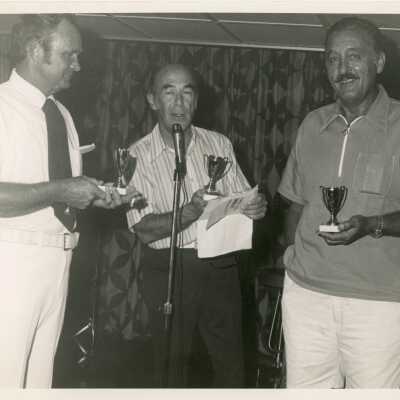 3 Unknown men standing at a microphone each holding a trophy