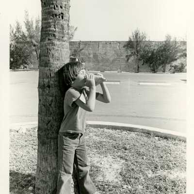 A child leaning against a tree blowing a conch
