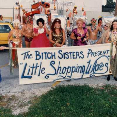 Unknown people dressed up holding a sign that reads The Bitch sisters present Little Shopping Whores.