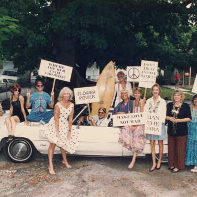 A white convertible with a group of unknown people some are standing near the car and some are sitting on it and in it.