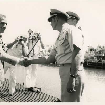 Men in uniform shaking an unknown man's hand with two others taking pictures
