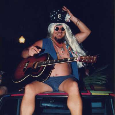 Unknown man dressed up sitting on the top of a car holding a guitar.