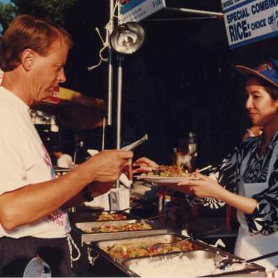 An unknown woman working a food booth, serving a plate of food to an unknown man.