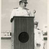 2 Men in uniform, one sitting and one standing at the podium