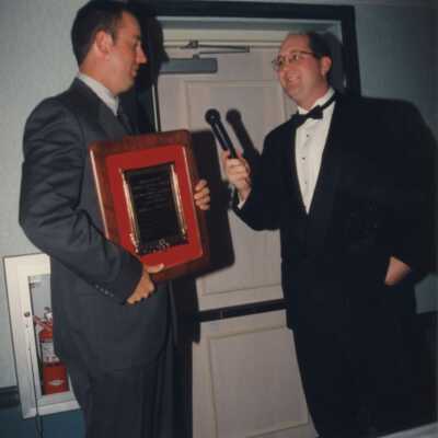 Two unknown men in suits. One has a microphone and the other has a plaque award.