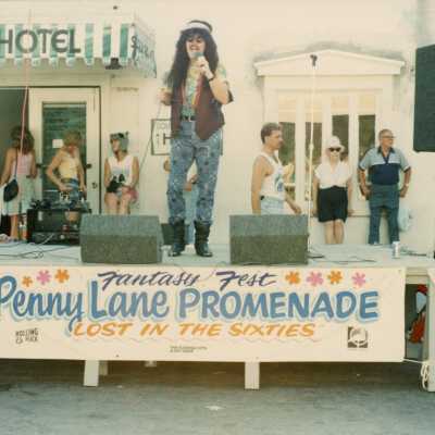 An unknown person dressed up on a stage. Sign reads Penny Lane Promenade.